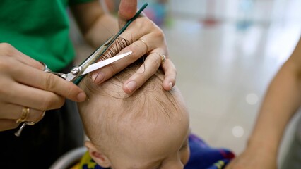 Cute blond boy in the barbershop getting his hair cut by the barber. Hands of a stylist with a comb. Baby's first haircut. Cute little boy at the professional children's hairdresser.