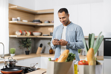 Attractive arab man preparing romantic dinner on home kitchen. Serious concentrated guy looking for good recipe at smart phone. Healthy food menu app. Cookbook mobile application. Technology and home