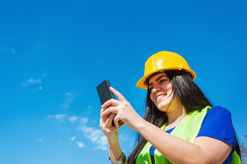 young woman worker with yellow safety helmet outdoors happy smiling checking her phone.