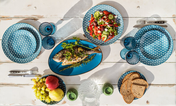 Gilt-head bream also known as Orata baked in the oven and served on a white wooden table with vegetable salad and fruits. Healthy seafood preparation and serving concept image.