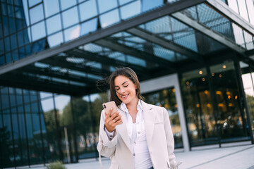 Young cheerful business woman working with a mobile phone in the street with office buildings in the background