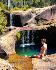 beautiful girl in pink bikini sits by natural rock pool with waterfalls at rainbow waterfall in...