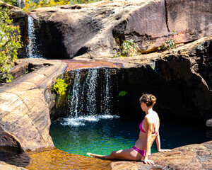 beautiful girl in pink bikini sits by natural rock pool with waterfalls at rainbow waterfall in blackdown tableland, holiday in australian outback, queensland