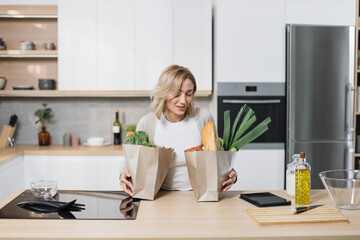 Beautiful young caucasian blond chef woman preparing to make delicious salad holding eco shopping bag with fresh vegetables and baguette in modern bright kitchen.