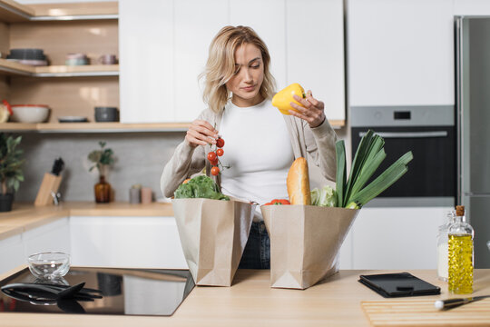 Young blond woman holding grocery shopping bag with vegetables standing in the modern light kitchen at home. Attractive female unpacking paper bag with fresh products. - Powered by Adobe