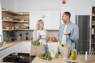 Arabian young man and woman in casual attire cooking together at home from healthy and fresh products. Happy family preparing fresh salad, meal on modern kitchen. Arab guy throwing tomatoes for fun.