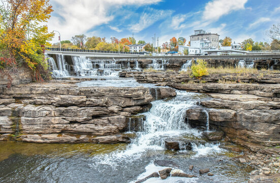 Waterfalls over large stone riverbed, more waterfalls and hydro dam buildings in background, fall foliage, sunny, blue sky, nobody