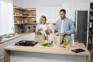Positive young people in love unpacking shopping back with fresh organic products. Caucasian family standing together on kitchen and enjoying domestic chores.