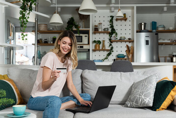 Fototapeta na wymiar Smiling happy woman paying online, using laptop, holding plastic credit card, sitting on couch at home, positive young female shopping, making secure internet payment, browsing banking service