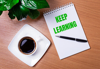 On the desktop there is a green plant, a white cup of coffee, a pen and an open notepad with the text KEEP LEARNING. Business concept. Close-up of the workplace.