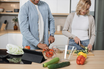 Cropped view of young attractive couple in love preparing salad from fresh vegetables. Handsome sporty man and blond charming woman cooking dinner together and having fun in a new modern apartment.