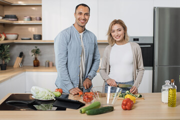 Young attractive couple in love preparing salad from fresh vegetables. Handsome sporty man and blond charming woman cooking dinner together and having fun looking at camera in a new modern apartment.