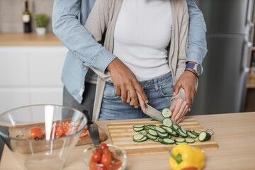 Cropped view of young attractive couple in love preparing salad from fresh vegetables. Handsome sporty man and blond charming woman cooking dinner together and having fun in a new modern apartment.