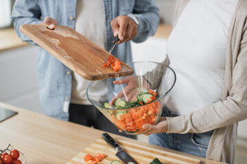 Close up of hands of couple preparing salad from fresh vegetables, man and woman dropping sliced tomatoes in glass bowl cooking dinner together in new modern kitchen at home