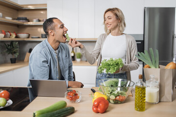 Cheerful married multinational couple using laptop while cooking healthy food in kitchen, blond young wife spouses having fun while feeding her husband with lettuce salad, preparing vegetable meal .