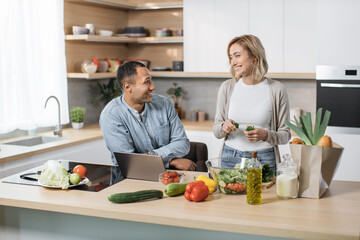 Attractive young multiethnic couple using laptop computer while preparing together healthy food diet vegetable salad at modern light kitchen at home.