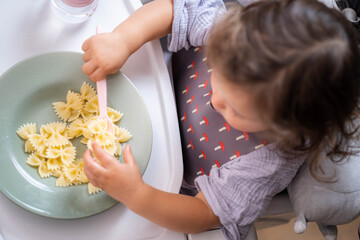 Top view portrait caucasian baby girl about 2 years old in bib eating pasta from plate sitting high...