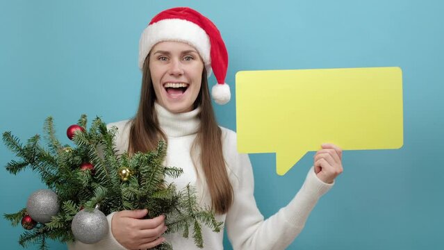 Happy young Santa female in Christmas hat holding bouquet of spruce branches and yellow empty blank Say cloud speech bubble, isolated on blue background. New Year celebration merry holiday concept