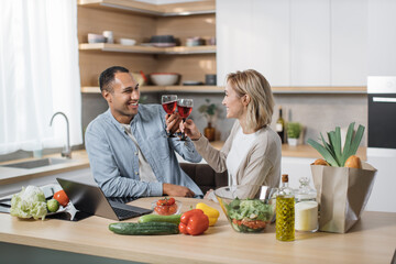 Happy multinational couple cooking dinner on the kitchen. Young people cook salad while drinking wine, toasting enjoy talking together. Beautiful multiethnic couple in love on romantic date at home.