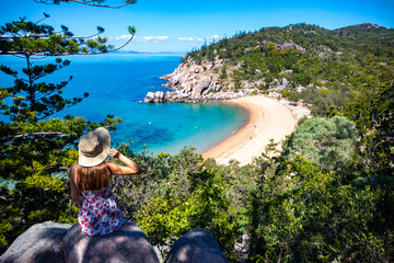 beautiful long-haired girl in dress and hat sits on top of mountain overlooking paradise beach on...