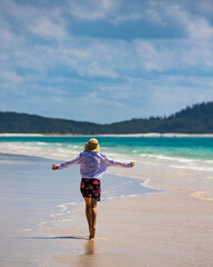 a beautiful girl in a dress, shirt and hat spreads her arms in joy as she walks along whitehaven beach; leisure on whistundays islands, queensland, australia; famous white sand dunes