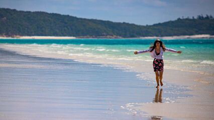 a beautiful girl in a dress, shirt and hat spreads her arms in joy as she walks along whitehaven beach; leisure on whistundays islands, queensland, australia; famous white sand dunes