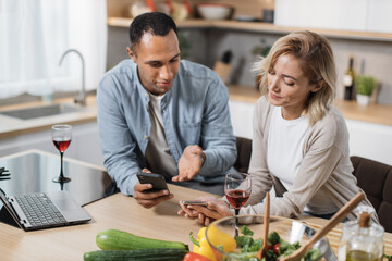 Close up view of multinational couple using cell phone while having breakfast at kitchen, positive...