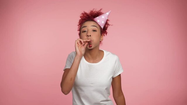 Happy African American redhead curly woman in birthday cap blowing whistle celebrating holiday. Joyful positive smiling female wearing white t-shirt on isolated pink background with copy space