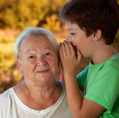 Boy whispering to grandma in summer outdoors garden, grandkid and grandmother lalking