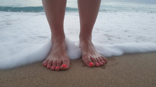 Feet of a young woman with painted nails on a sandy beach, washed by foam and waves of the ocean