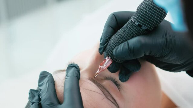 Cosmetologist creating new shape of model eyebrows using ink machine during permanent makeup process