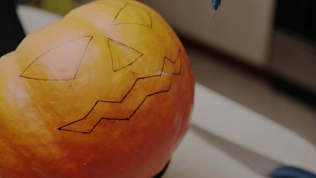 A woman draws a scary face on an orange pumpkin with a black marker in the kitchen. Halloween holiday 