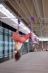 Sporty woman doing aero yoga in hammock. Brunette girl in sportswear exercising aerial yoga on terrace. Windows in background