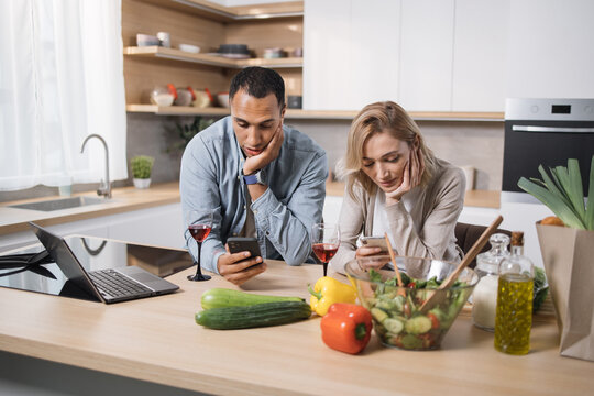 Tired, emotionless multinational couple eating salad using smartphone checking photos on social media, network in their modern light kitchen at home.
