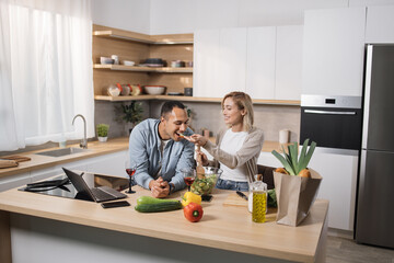 Cheerful married multinational couple using laptop while cooking healthy food in kitchen, blond...