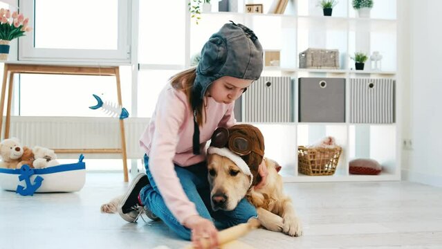 Little girl and golden retriever dog wearing pilot hats playing in the room with wooden plane