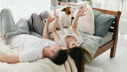 Young couple lying in the bed in the room with sunlight and peting cute small dog. Attractive woman and handsome man spend morning time together in the bed and smiling in their bedroom with pet doggy