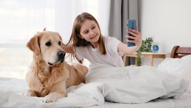 Beautiful preteen little girl sitting in the bed with golden retriever dog with smartphone. Cute smiling kid with pet and cell phone.