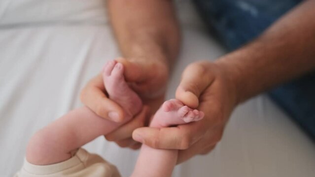 Father holding tiny sweet feet of newborn baby in his hands and petting them. Dad of adorable infant cares about his child at home