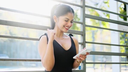 Overjoyed attractive indian woman celebrate online win, success hearing great news, feel amazed while her workout near the stadium. Closeup of girl with good news on her phone.