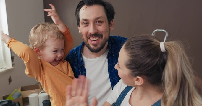 The man is holding two-year-old son, while the wife is holding camera and having video call with family. The boy in orange sweater is having fun, parents are talking, smiling and showing new home.