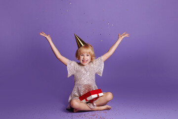 A child in a festive cap and a sparkling dress sits on a purple background and throws sparkles over his head. Holiday concept, congratulations. Place for your text.