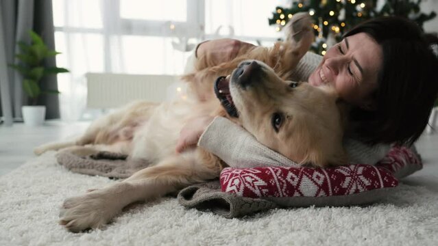 Girl petting golden retriever dog on floor in Christmas time at home with New Year decoration and smiling. Young woman with pet doggy together in Xmas