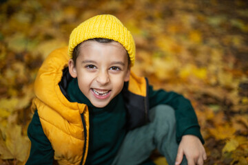 portrait of a fashionable child boy autumn sitting on a trail in orange leaves in the afternoon at a green fence on the street.