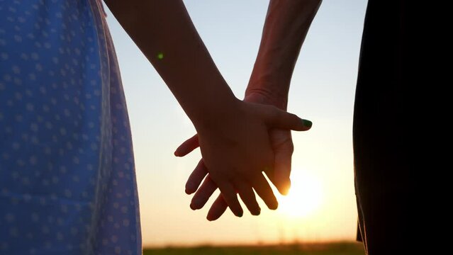 Close up of brother and sister holding hands against sunset in field. Caring and support in loving family. Happy childhood. Walk together in outdoor summer nature in evening. Rear view two children.