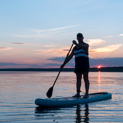 A man in shorts and a T-shirt on a SUP board against the backdrop of the sunset sky in the lake.