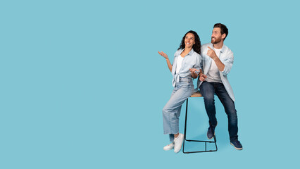 Fototapeta premium Surprised happy millennial caucasian husband and arab wife sitting on chair together, show fingers to empty space