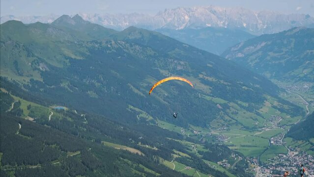 Man parachuting paragliding with a bright orange parachute slowly flying in the air sky in between green mountains with beautiful panorama view of the alps. Skydiving extreme action sport in summer. 