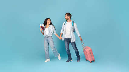 Cheerful millennial caucasian man and arab female with suitcase and air tickets holding hands