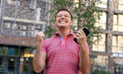 Excited millennial guy receives incredible news on phone, raises his hand in victorious gesture and...